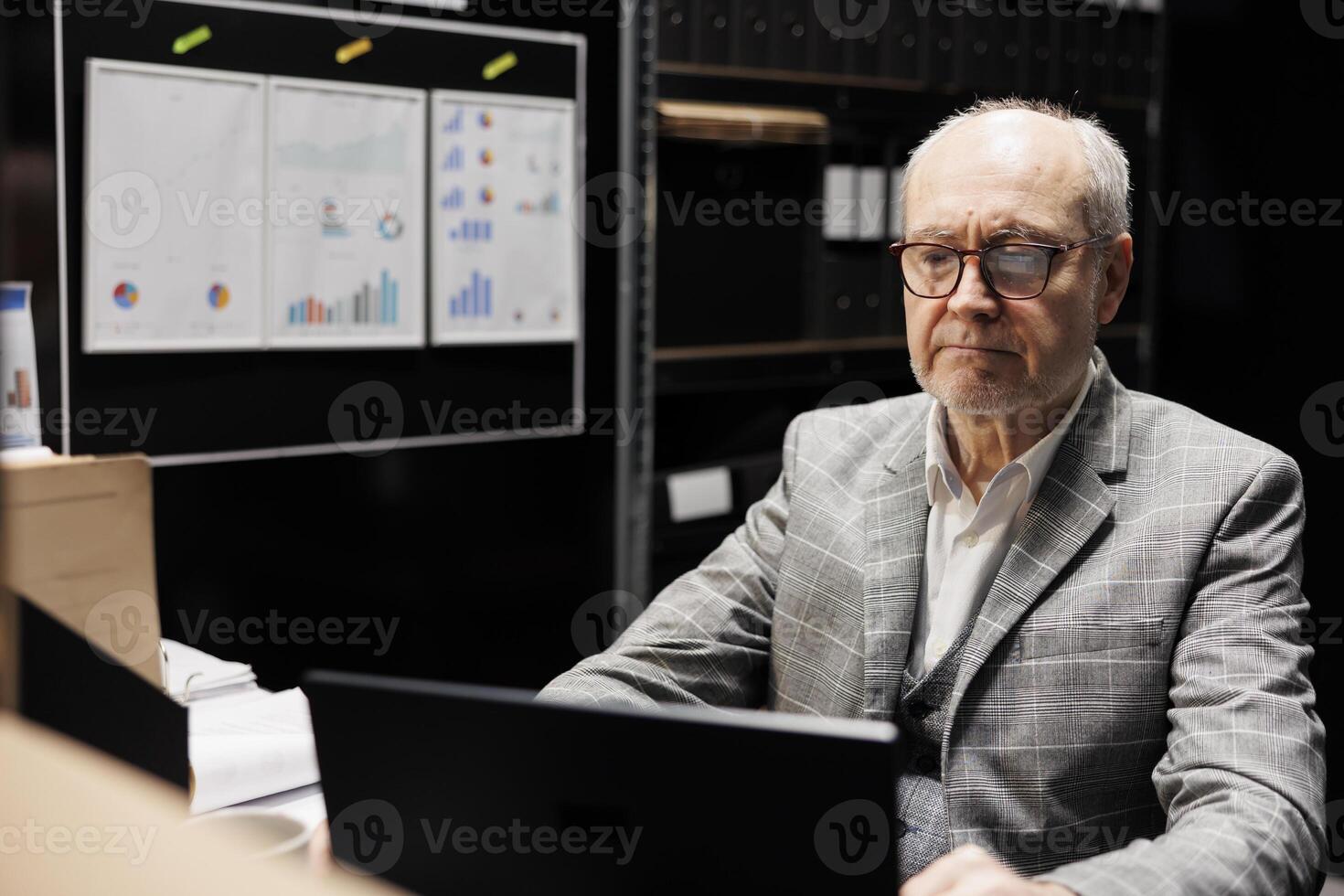 Old businessman employee inserting accountancy budget plan data on laptop. Senior bureaucratic executive in storage room office filled with graphic charts reports and document folders photo