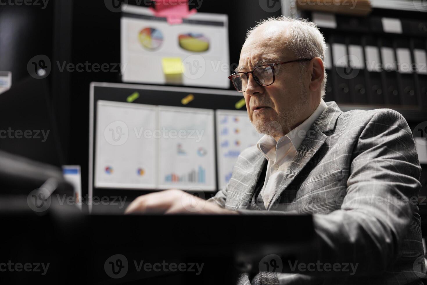 Old business manager executive sitting at office desk before consulting meeting, inspecting quarterly company results analysis data paperwork on laptop. Bureaucratic workplace filled with flowcharts photo
