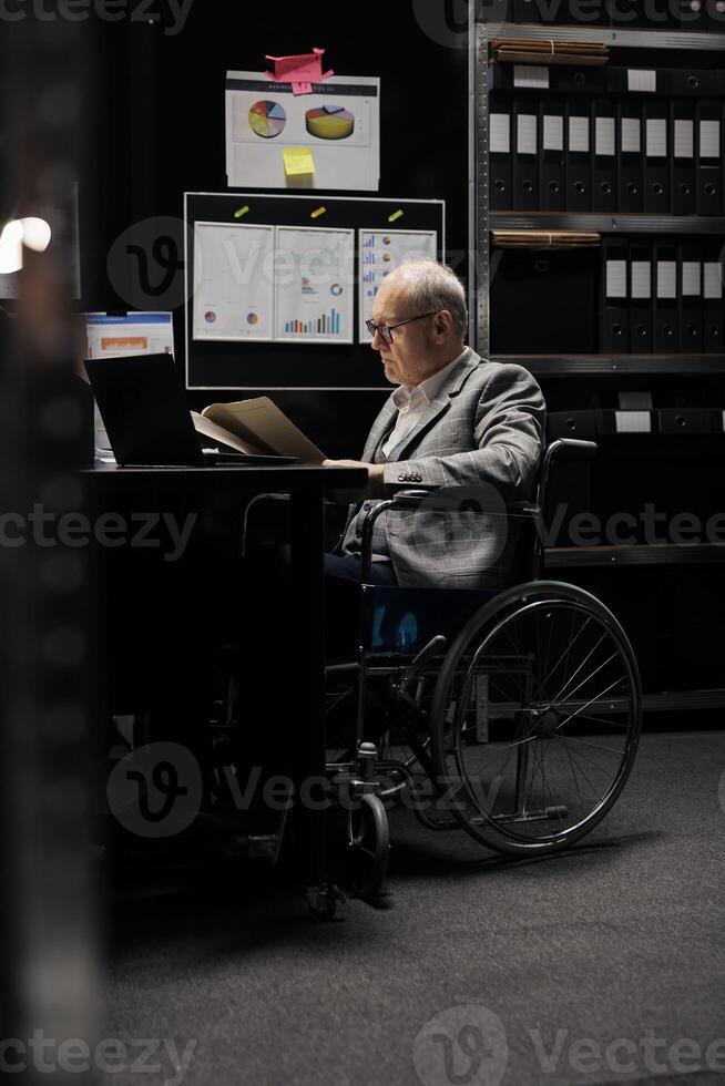 Senior wheelchair user checking annual profit revenue paperwork before business consulting meeting. Executive with paraplegia doing objective completion measurements in file cabinet office photo