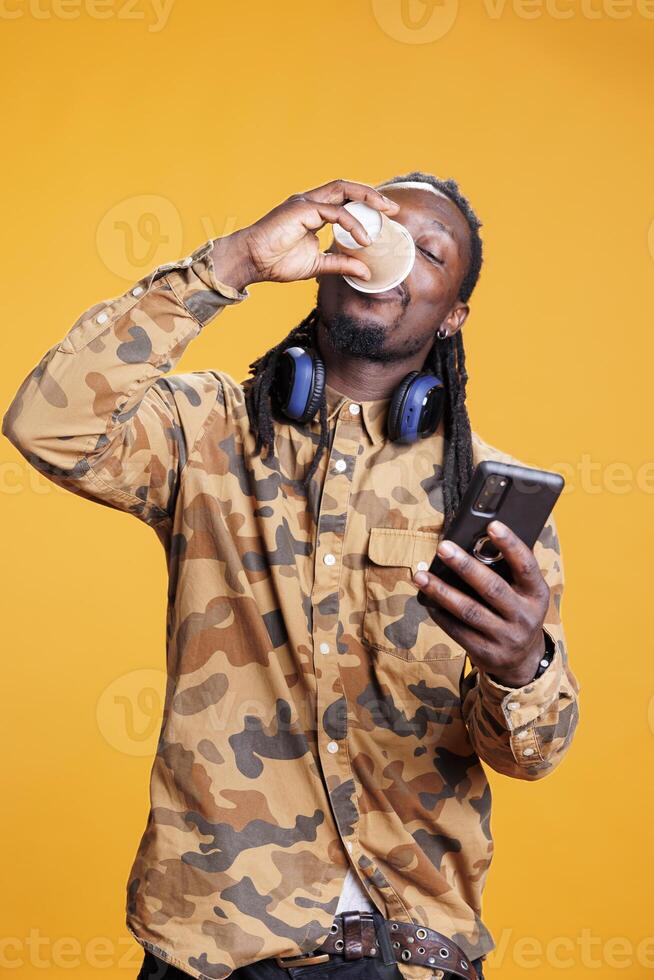 African american adult drinking hot coffee, holding smarphone scrolling to social media during lesiure time in studio. Cheerful man searching information on internet using mobile phone photo