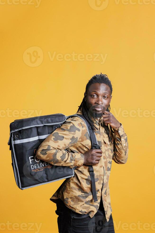 Cheerful courier holding food delivery thermal backpack, standing in studio over yellow background. African american restaurant worker deliverying fast food order to customers during lunch time photo