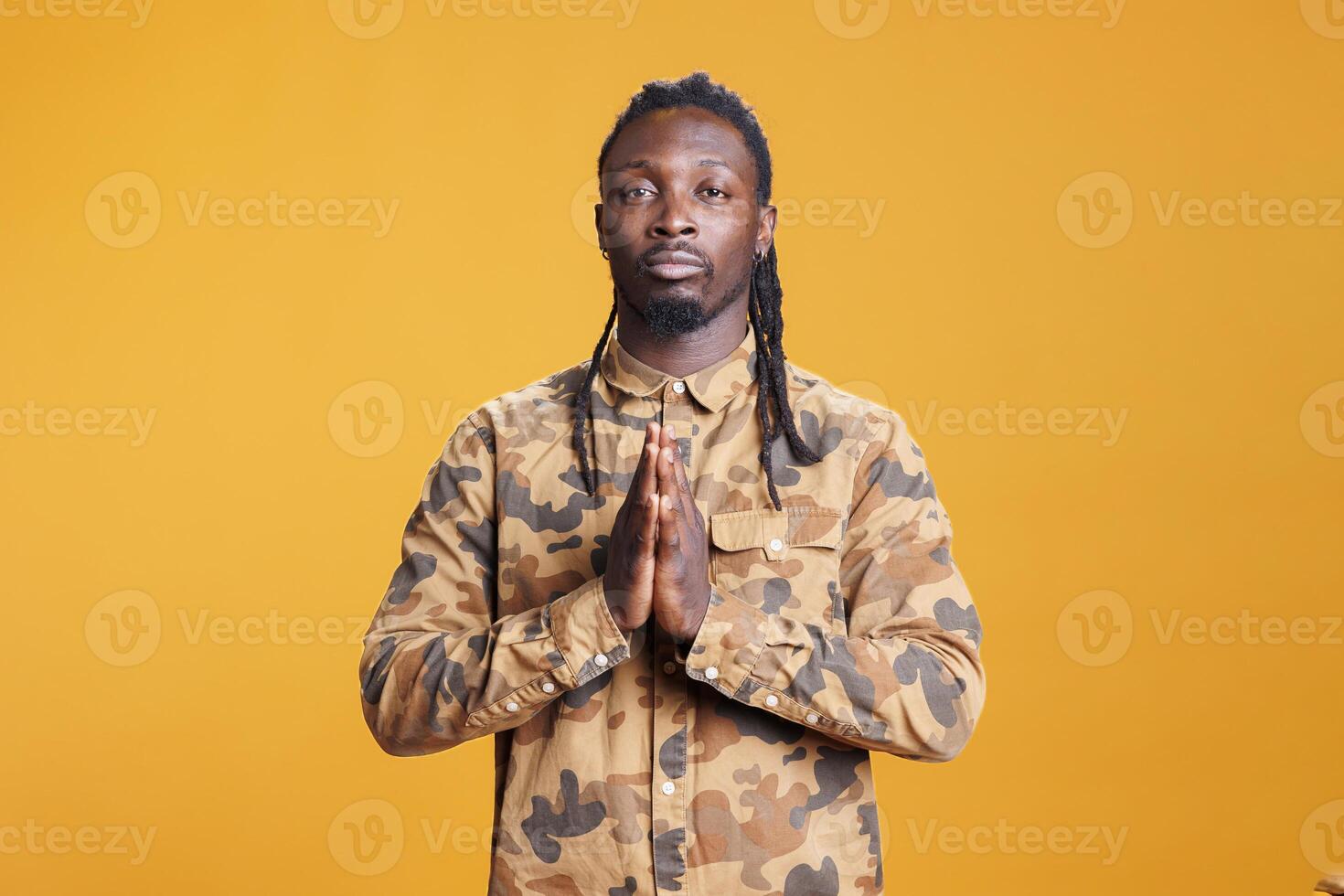 African american man doing prayer hands symbol in studio, showing religious spiritual gesture over yellow background. Hopeful young adult praying to jesus for peace and forgiveness. photo