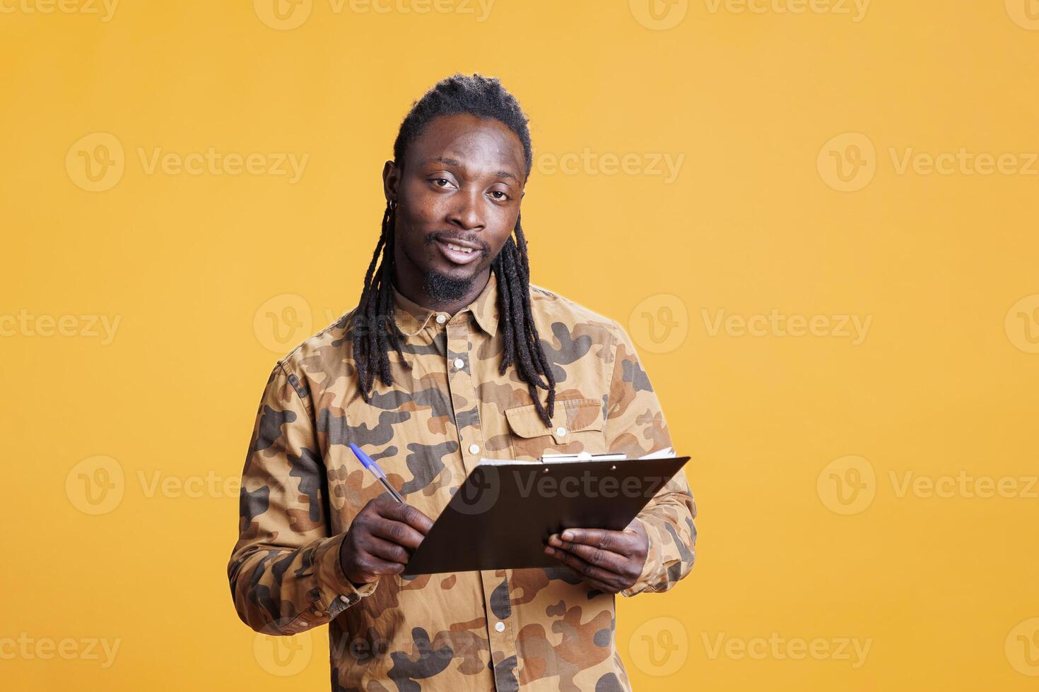 Young adult holding clipboard documentation, writing business job schedule on papers in studio over yellow background. African american man analyzing checklist, signing agreement files photo