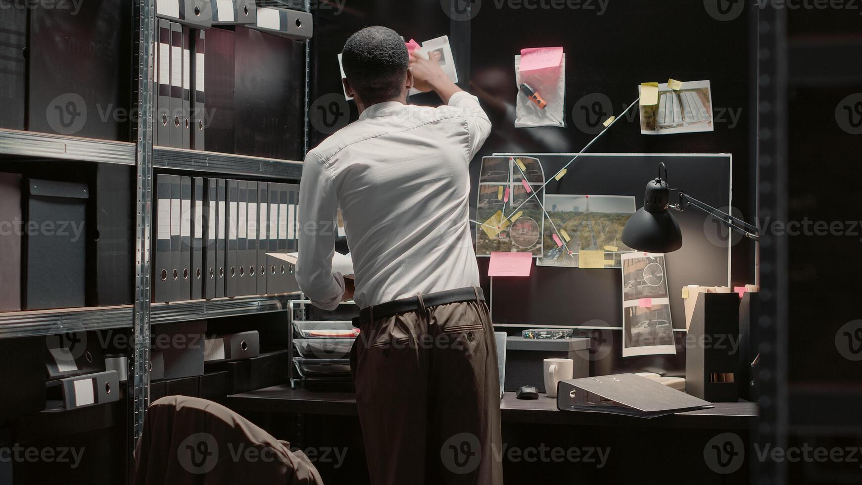 Detective sitting at desk analyzing evidence map on wall, examining law enforcement archive to solve crime. Male investigator studying clues and case files photos on detective board.