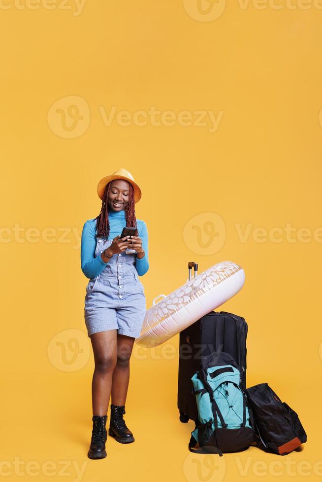 Young tourist checking smartphone app, posing with trolley bags and inflatable in studio. Female model feeling happy on inernational vacation, using mobile phone online browser. photo