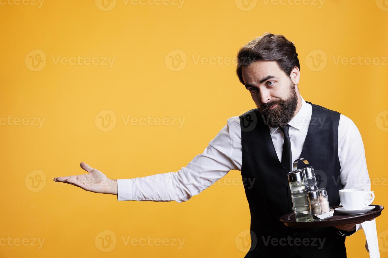 Friendly gracious waiter makes way for people to enter restaurant, providing great service and amazing food at five star place. Young adult with suit and tie posing against yellow background. photo