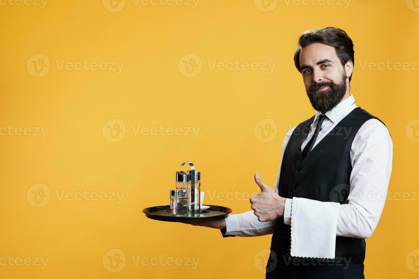 Smiling butler does like sign in studio to express positivity and approval with thumbs up symbol. Elegant happy restaurant worker presents agreement gesture against yellow backdrop. photo