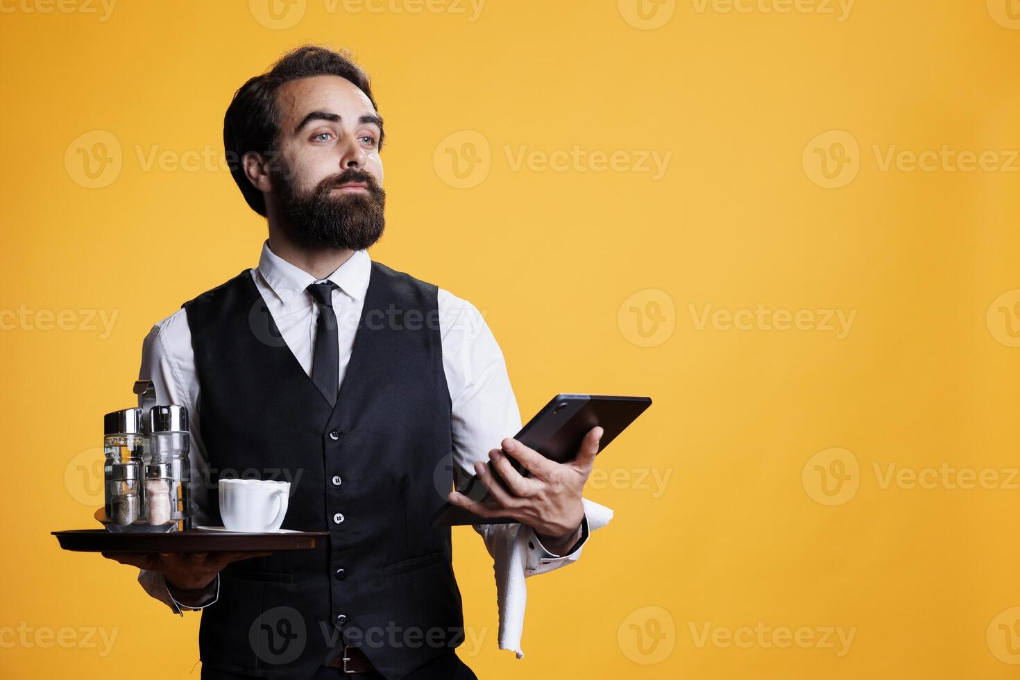 Luxury butler checking list of reservations on tablet, carrying tray with food and drinks. Elegant restaurant waiter preparing to give bill or tab to customers, using modern device. photo