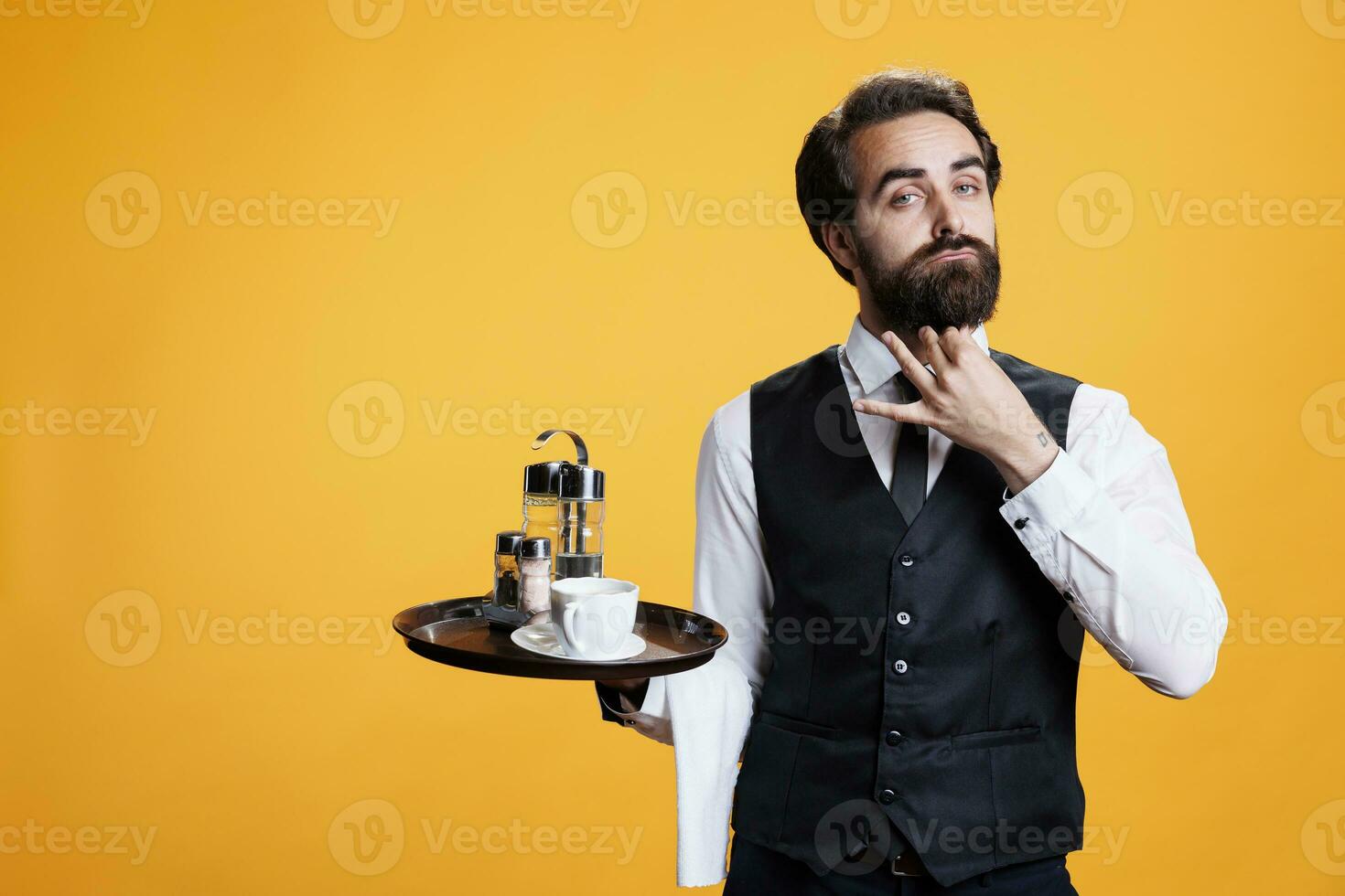 Stylish waiter posing with food tray against yellow background, preparing to serve coffee cup or meal. Confident elegant restaurant employee in front of camera, feeling determined. photo