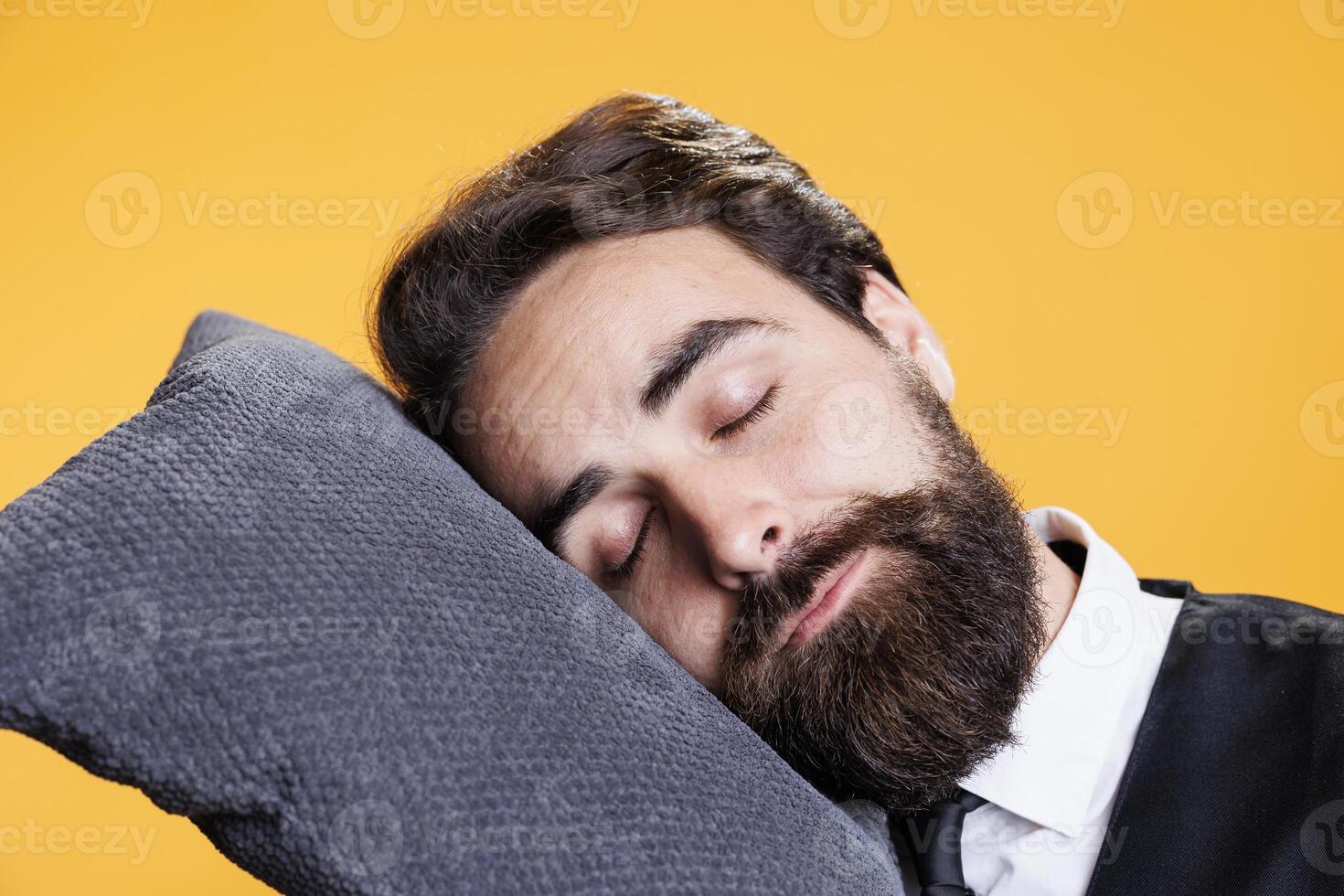 Tired waiter falling asleep on camera, taking a nap while he in standing against yellow background. Young sleepy person being overworked with burnout, falling asleep on pillow in studio. photo