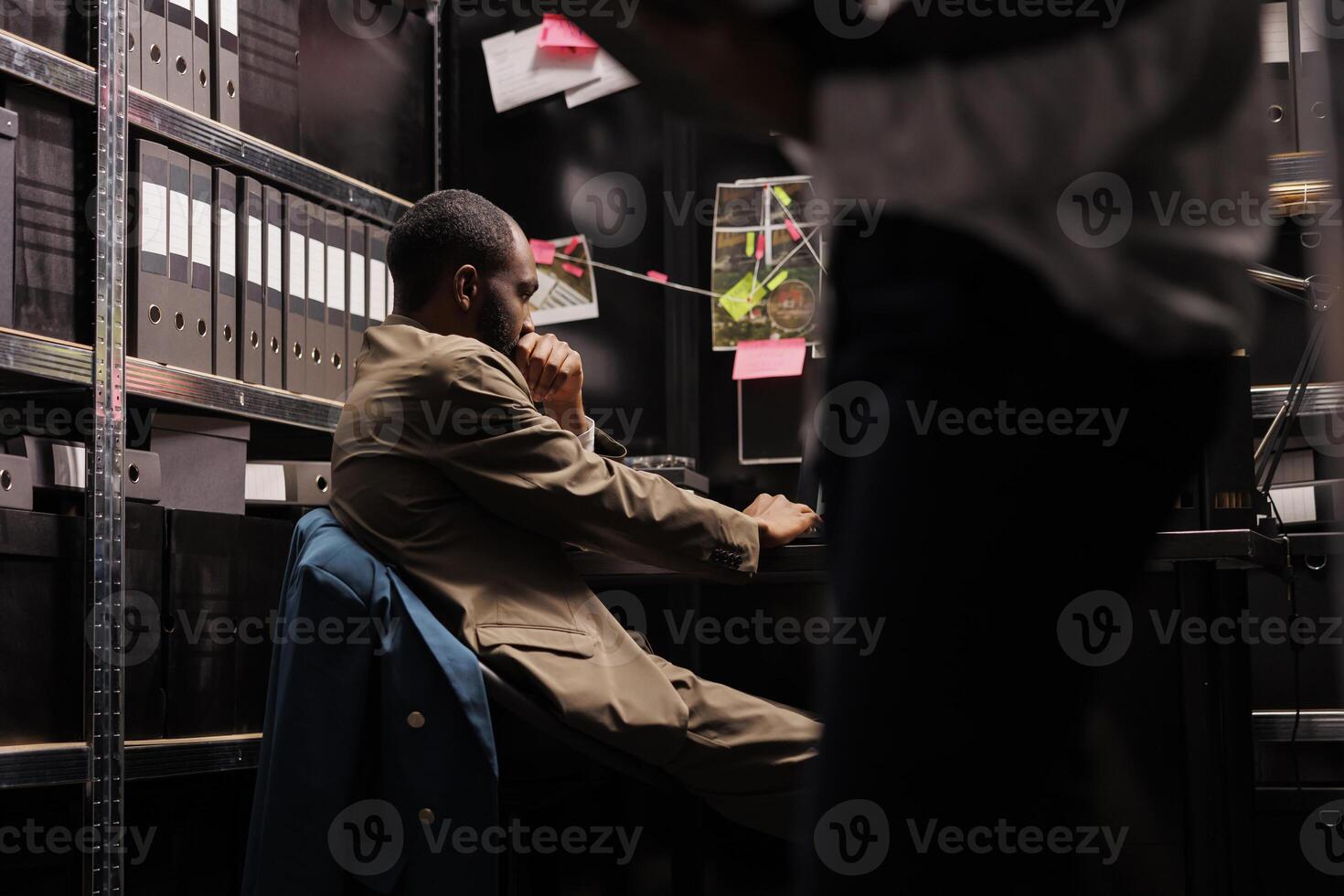 africano americano detective trabajando a lugar de trabajo escritorio en agencia oficina. preocupado investigador lluvia de ideas, pensando acerca de resolviendo crimen mientras sentado a mesa a noche hora foto