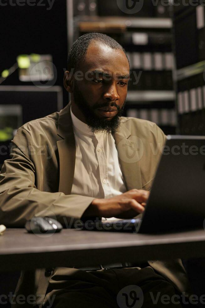 Concentrated private detective checking evidence using laptop while working overtime in dark agency office. Investigator reading police records on computer, searching insight for solving crime photo