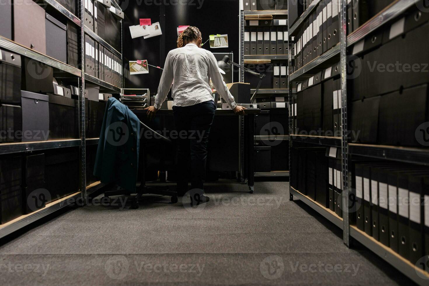 Police detective standing near desk and analyzing clues board in dark office late. African american woman police investigator working overtime and studying crime case evidence photo