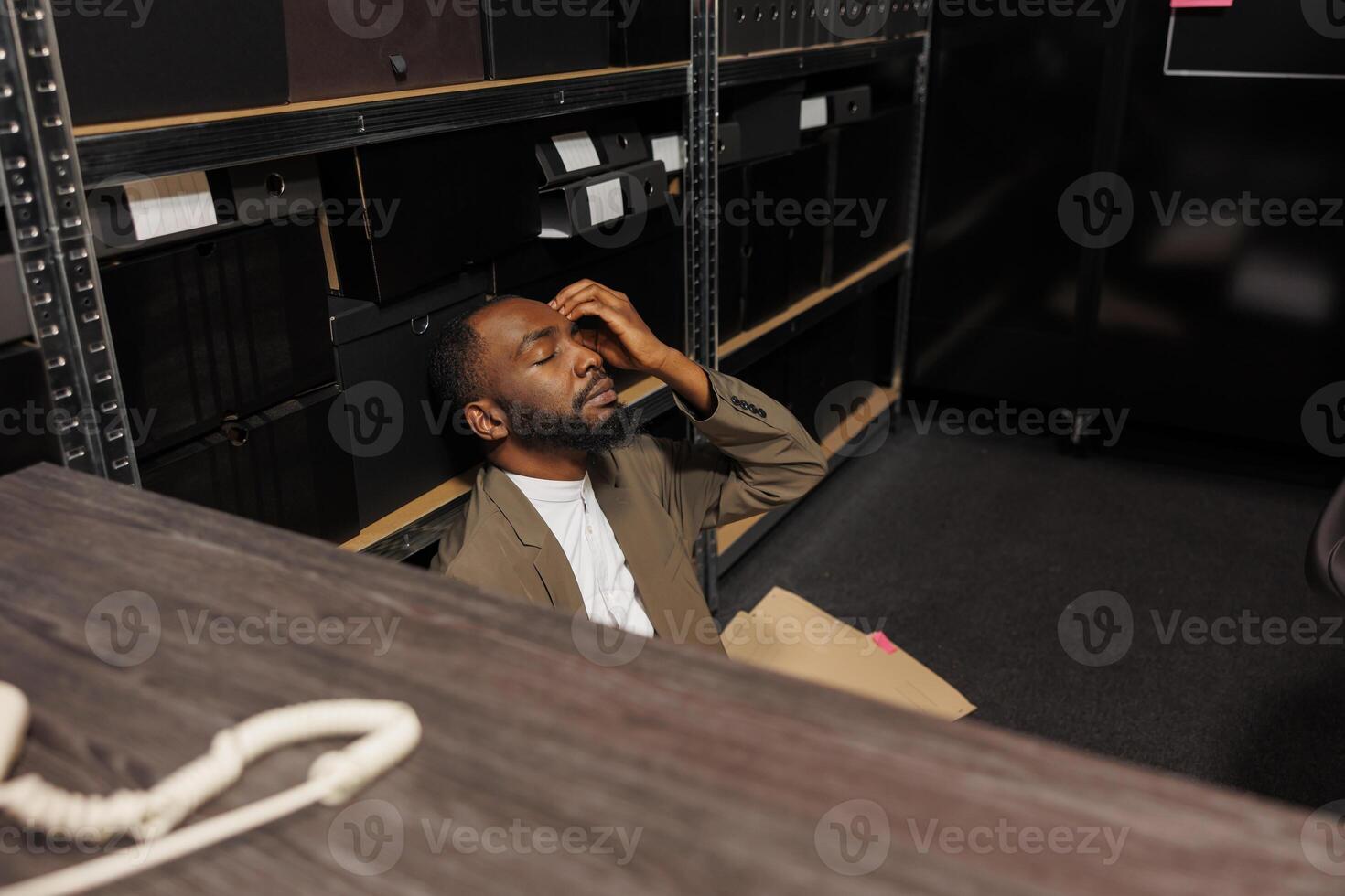 Stressed cop sitting on floor closing eyes and solving crime in police office at night time. Tired sleepy african american investigator conducting investigation and analyzing clues photo