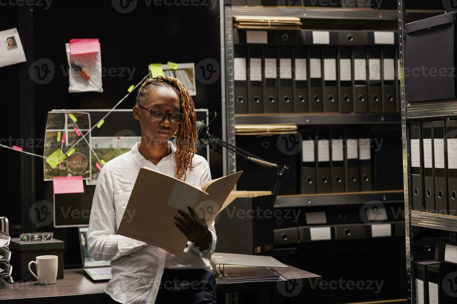 Serious african american woman detective studying crime matter file. Police investigator holding folder with criminal investigation information in office room at night time photo