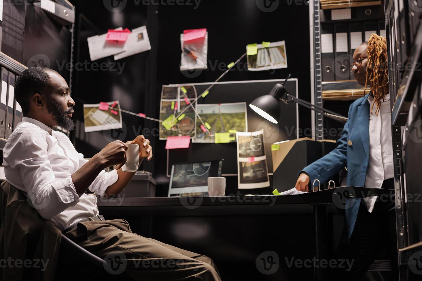 Detectives team conducting crime case investigation together, drinking coffee and reading files. African american man and woman cops teamwork cooperation in police investigator room at night time photo
