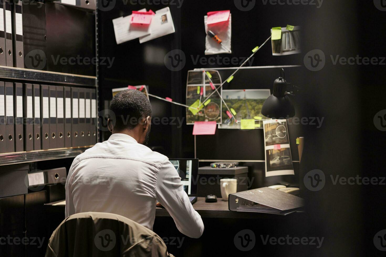 Police investigator working overtime analyzing evidence to solve crime. Criminalist sitting at desk, checking suspect photos on laptop and studying clues on detective board