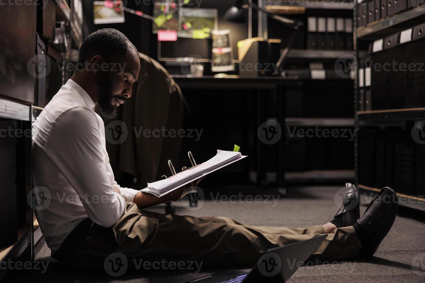 Detective sitting on floor and studying investigation report in dark office. African american policeman holding folder with crime case and analyzing information at night time photo