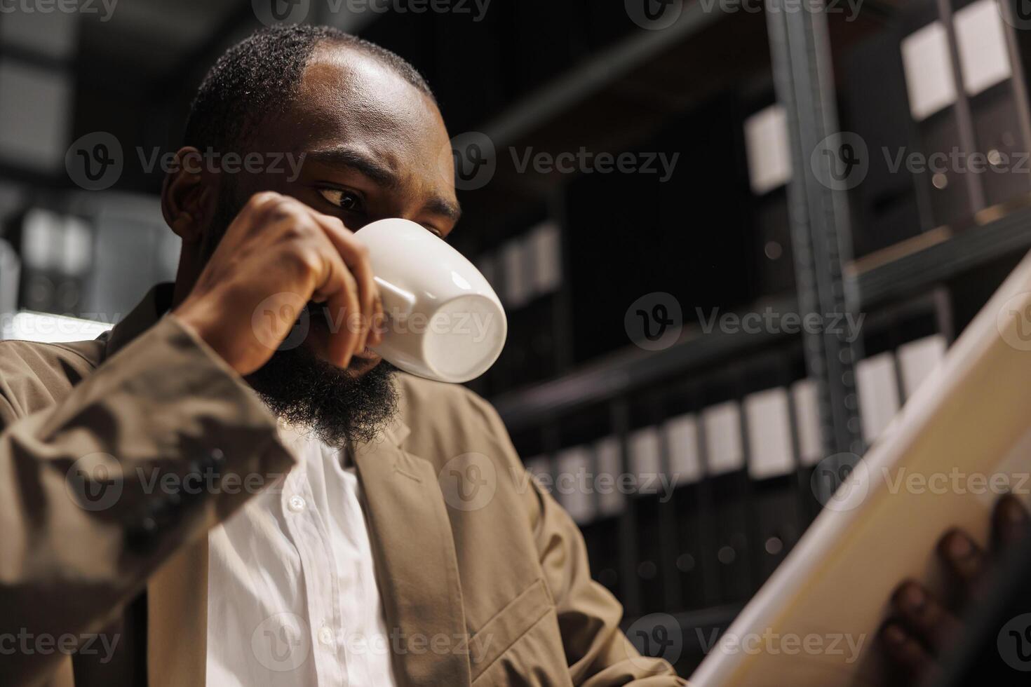 Private investigator drinking coffee while examining police documentation. African american man detective working late and reading crime matter file in agency office at night time photo