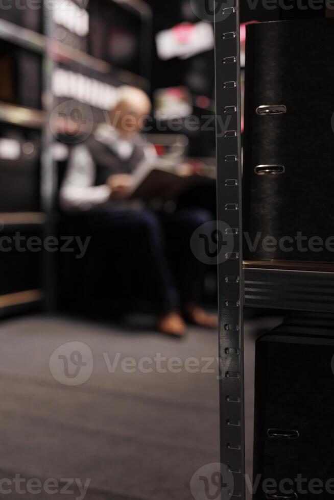 Selectiv focus of metallic storage full with confidential files, in background elderly private detective analyzing crime scene evidence. Old police officer working overtime at criminal investigations photo