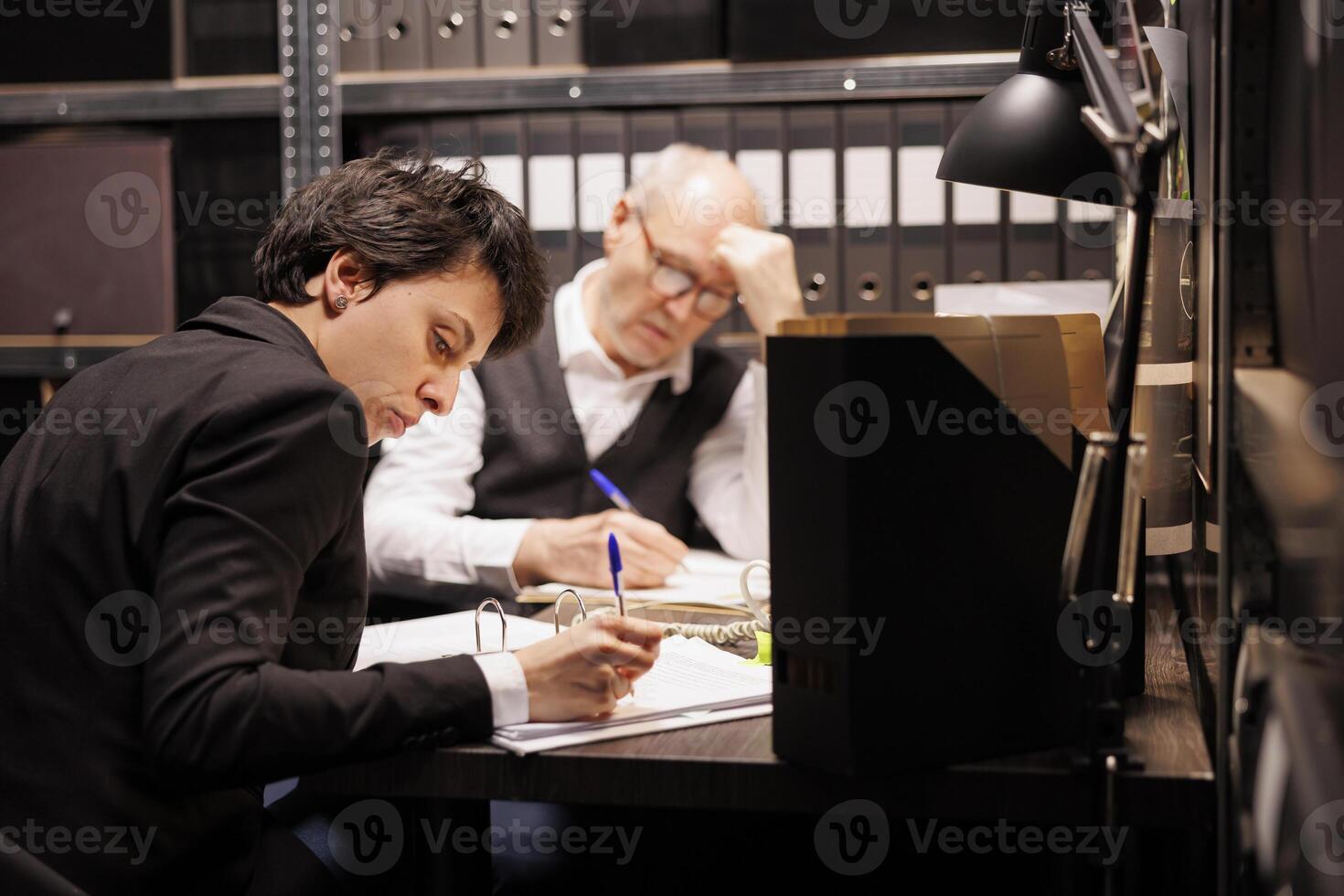 Inspectors working overtime at criminal investigations in arhive room, analyzing mysterious crime case. Elderly police officer checking federal evidence while investigator writing criminology report photo