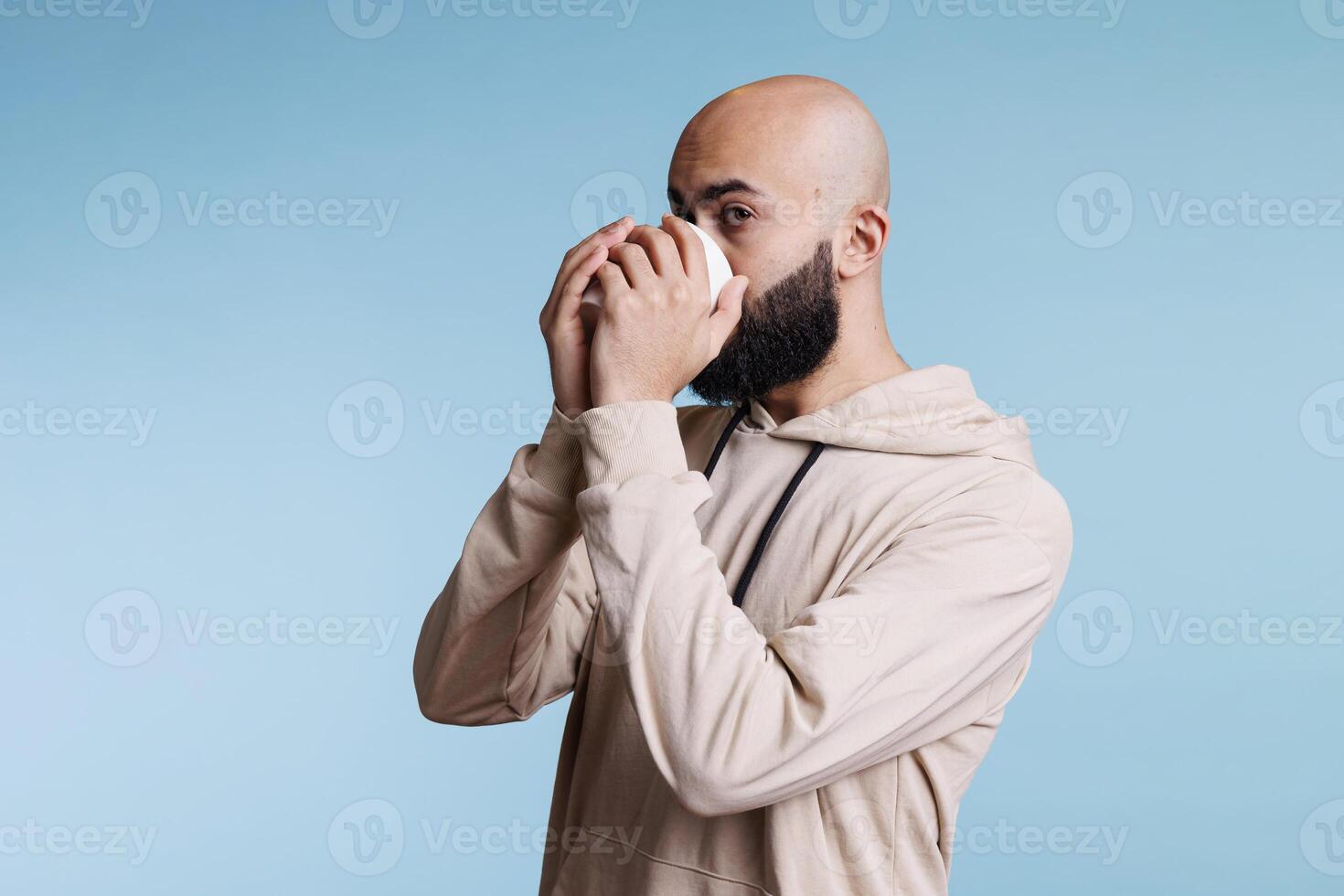 Young bald arab man enjoying morning cup of tea with relaxed facial expression. Person taking sip of coffee, drinking hot beverage from white mug on studio blue background photo