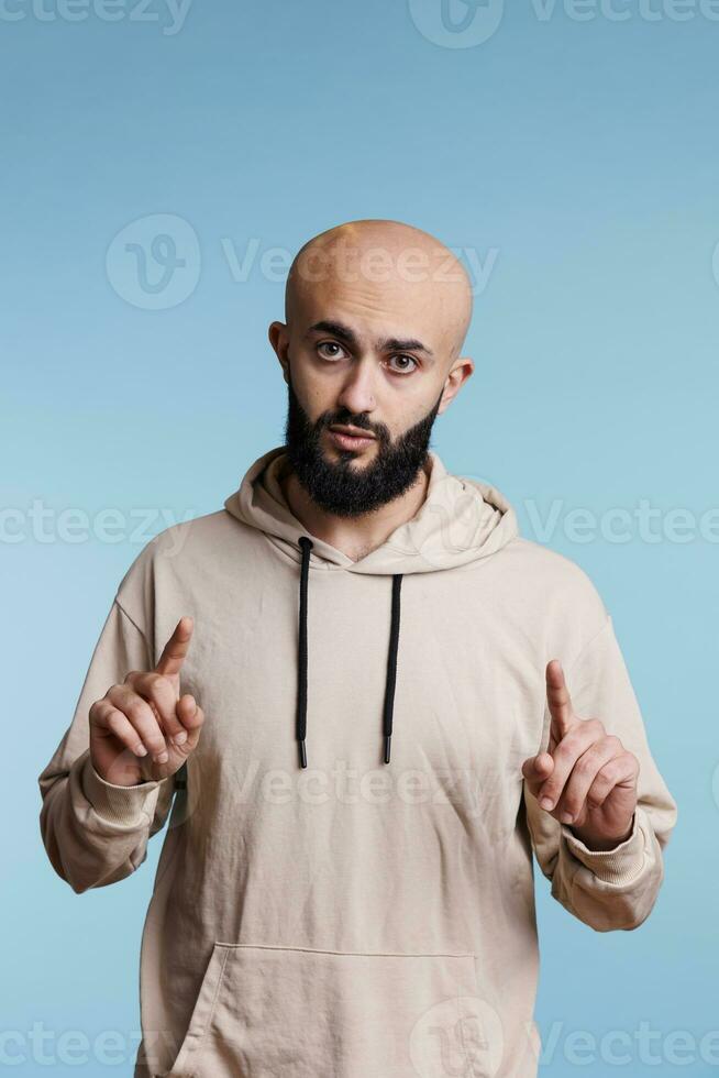 Young arab man pointing at camera studio portrait, interrupting with question. Handsome arabian person wearing casual clothes lifting two index fingers and looking at camera photo