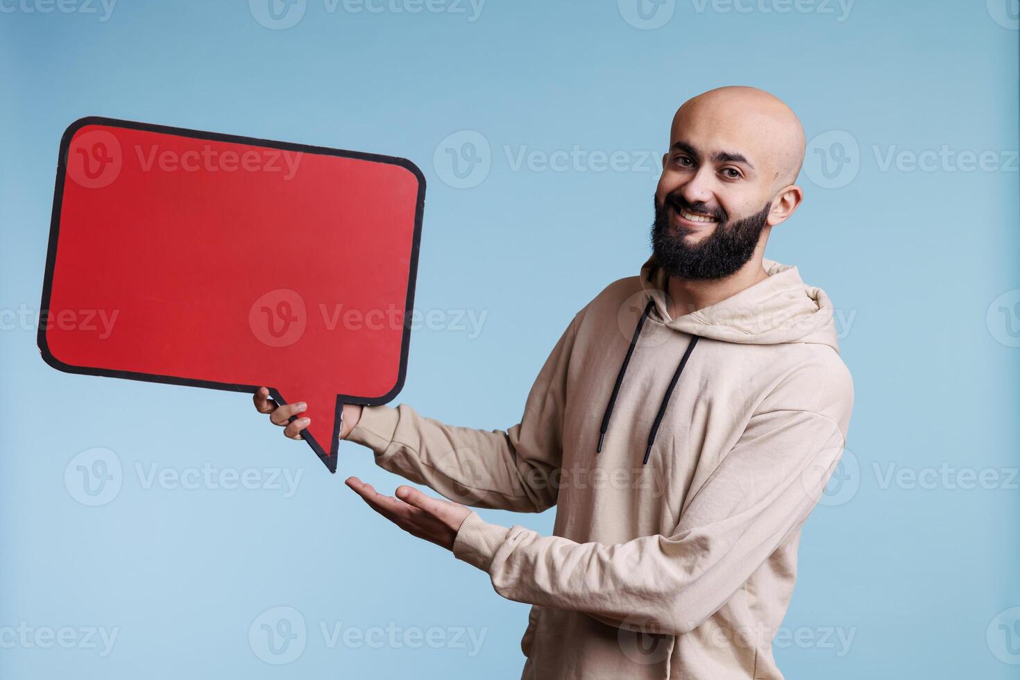 Cheerful arab man promoting new product with comic balloon red banner portrait. Smiling person holding empty dialog bubble and looking at camera with happy facial expression photo
