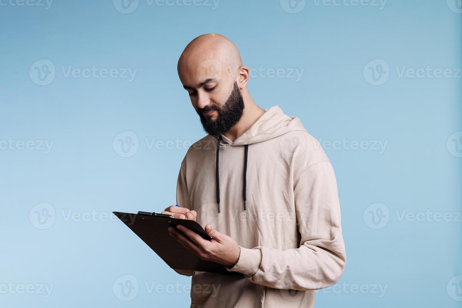 Concentrated arab man planning and writing checklist while holding clipboard. Young focused person wearing casual hoodie checking items in list and taking notes in studio photo
