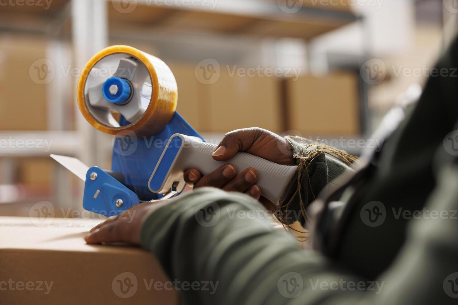 African american employee preparing customers orders, putting adhesive tape on cardboard box before shipping packages. Stockroom supervisor working at goods inventory in storage rooom. Close up photo