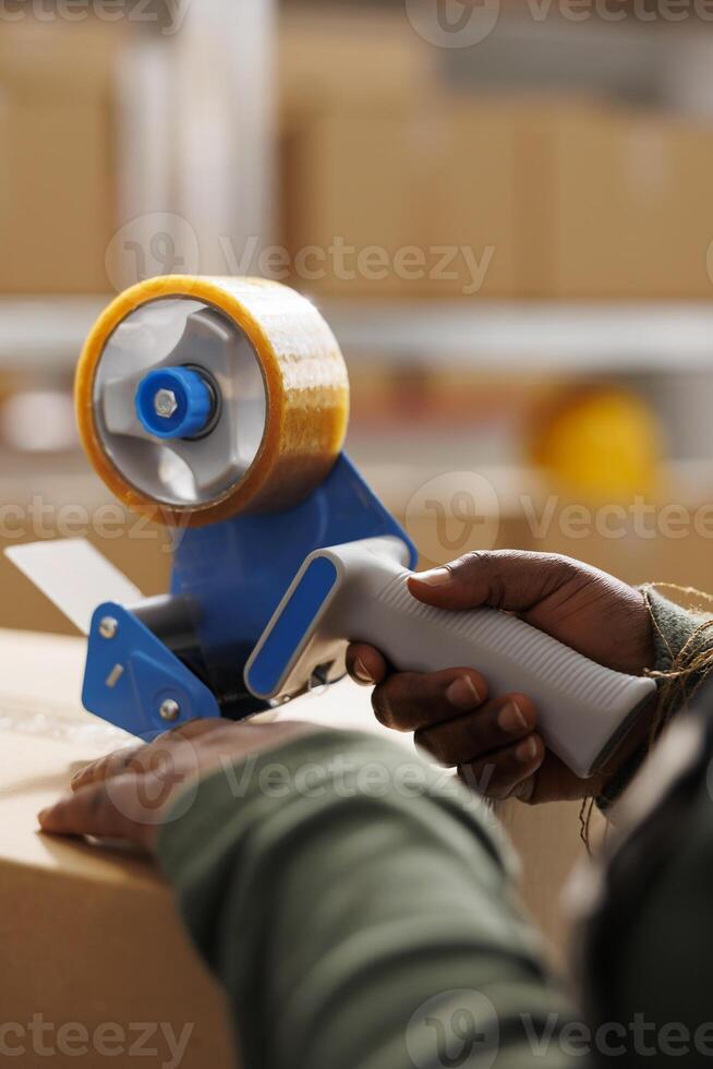 Warehouse employee putting adhesive tape on carton boxes, preparing customers orders in storage room. African american worker checking products before shipping packages. Close up shot photo