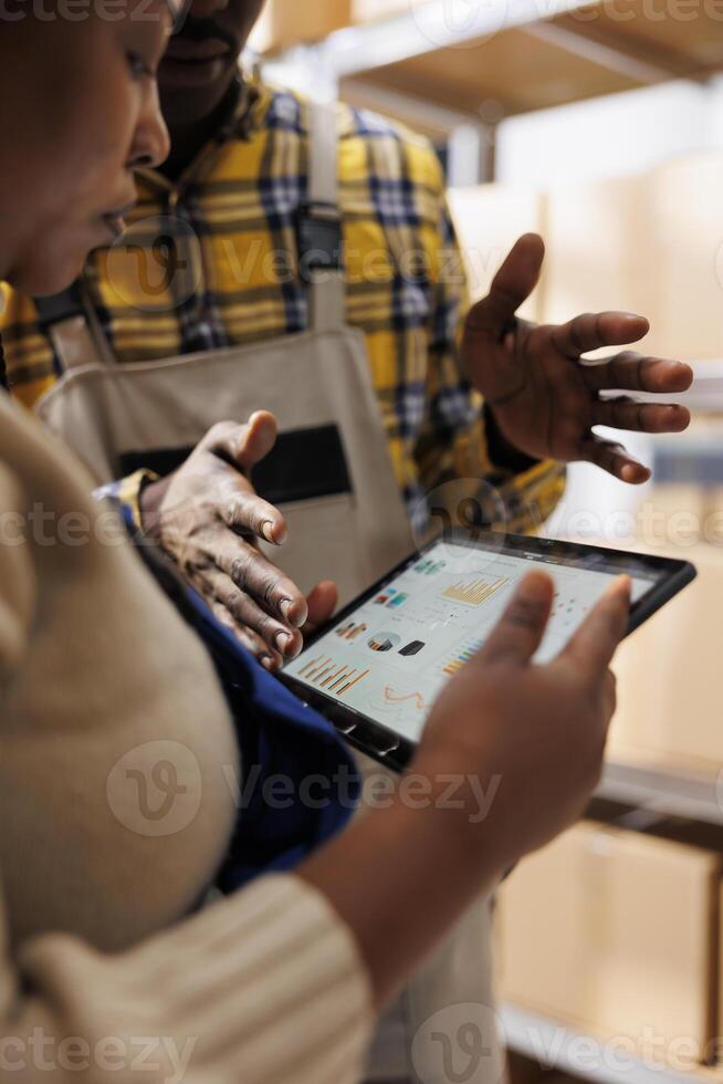 Inventory analyst explaining warehouse employee data on tablet screen. African american industrial storehouse manager holding portable device and analyzing stock supply analytics photo