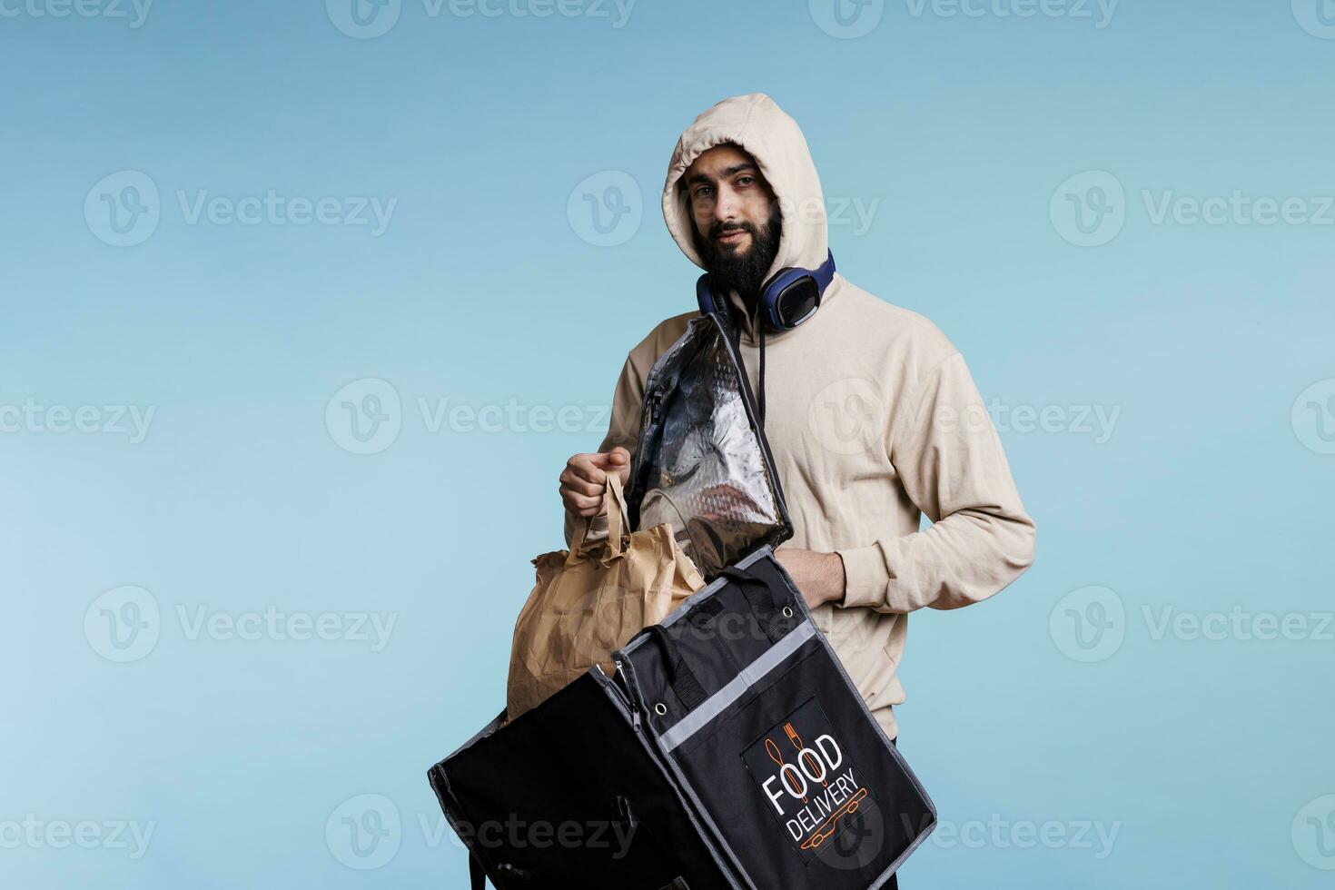 Young arab man taking paper bag with food from backpack and looking at camera. Deliveryman in hood holding thermal bag and package with takeout lunch studio portrait on blue background photo