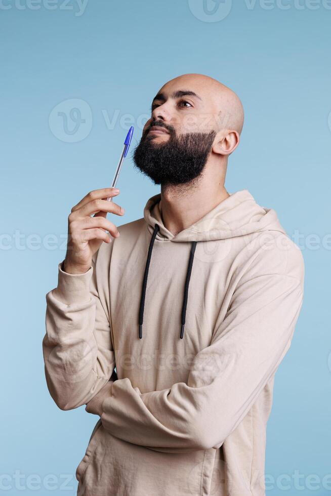 Thoughtful arab man holding pen and looking upwards in contemplation. Pensive young adult bald bearded person standing in thinking pose with questioned unsure facial expression photo