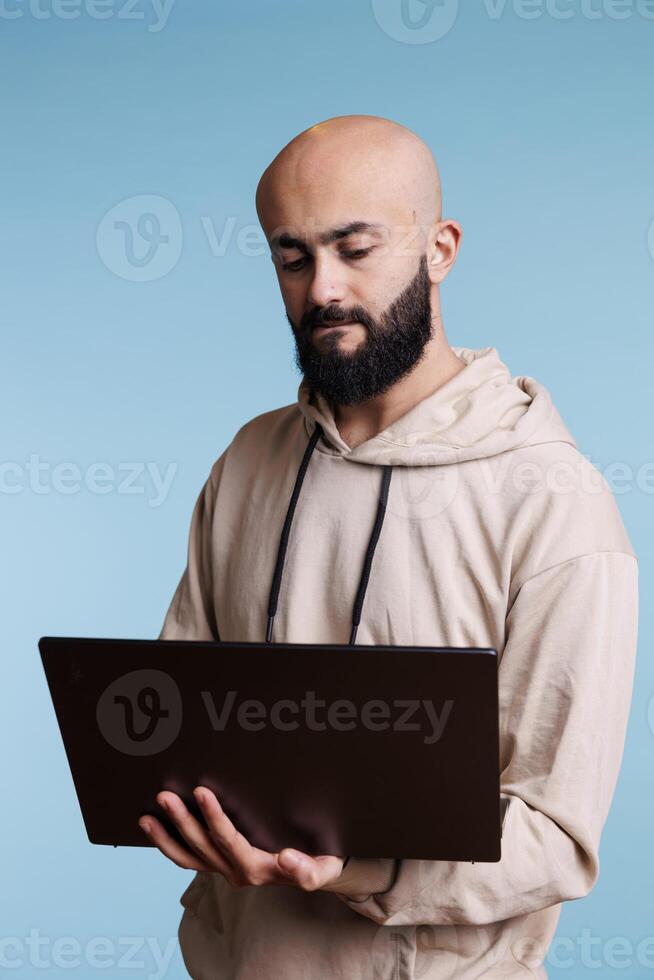 Concentrated arab man reading business report on laptop and typing online message. Focused young person holding portable computer, using software apps and browsing internet photo