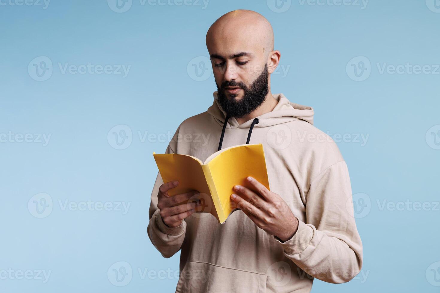 Young thoughtful focused arab man reading novel from book with yellow cover. Concentrated bald bearded person holding open softcover textbook and studying with pensive expression photo