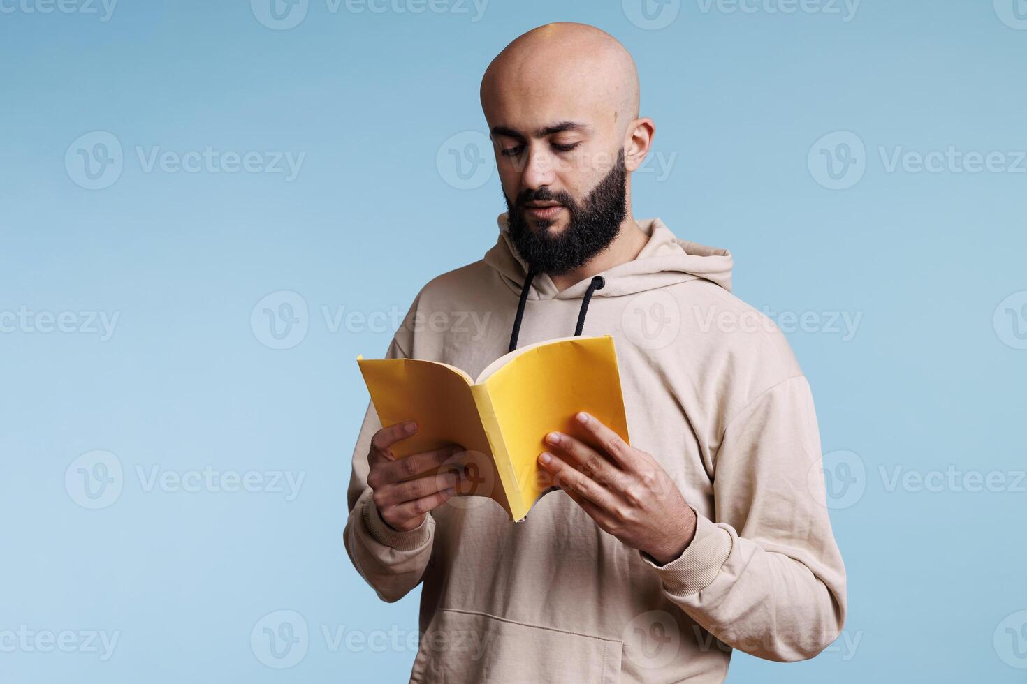 joven pensativo enfocado árabe hombre leyendo novela desde libro con amarillo cubrir. concentrado calvo barbado persona participación abierto tapa blanda libro de texto y estudiando con pensativo expresión foto