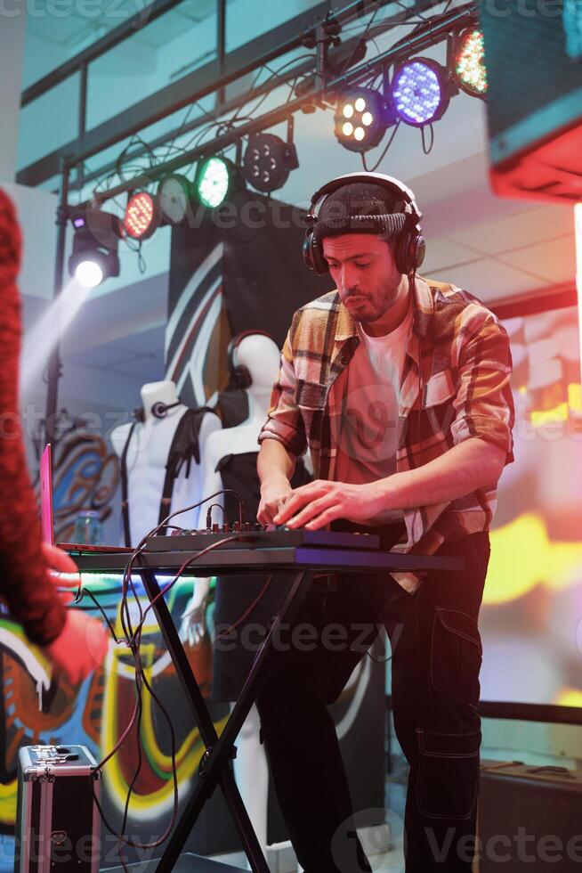 Performer playing techno music at discotheque in nightclub. Young man dj in headphones using digital controller for mixing sound on stage with spotlights at party in club photo