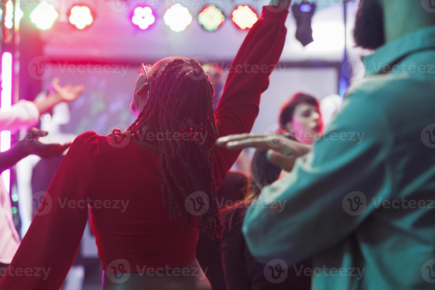 African american woman in red blouse jumping and dancing in crowded nighclub. Young clubbers having fun and partying on dancefloor with spotlights at discotheque in dark club photo