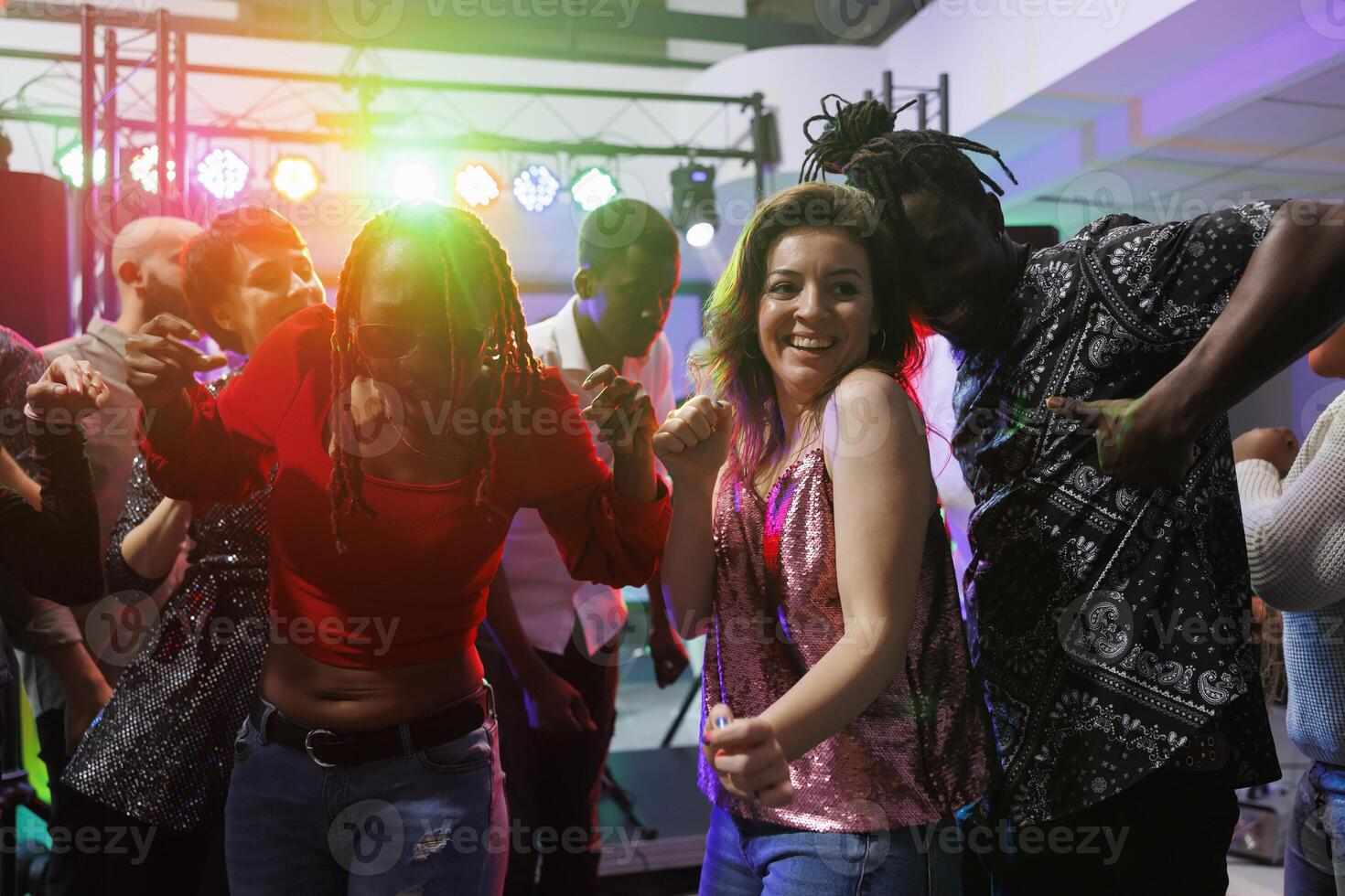 Smiling woman dancing with boyfriend on nightclub dancefloor illuminated with spotlights. Cheerful passionate diverse couple having fun and moving to music rhythm in crowded club photo