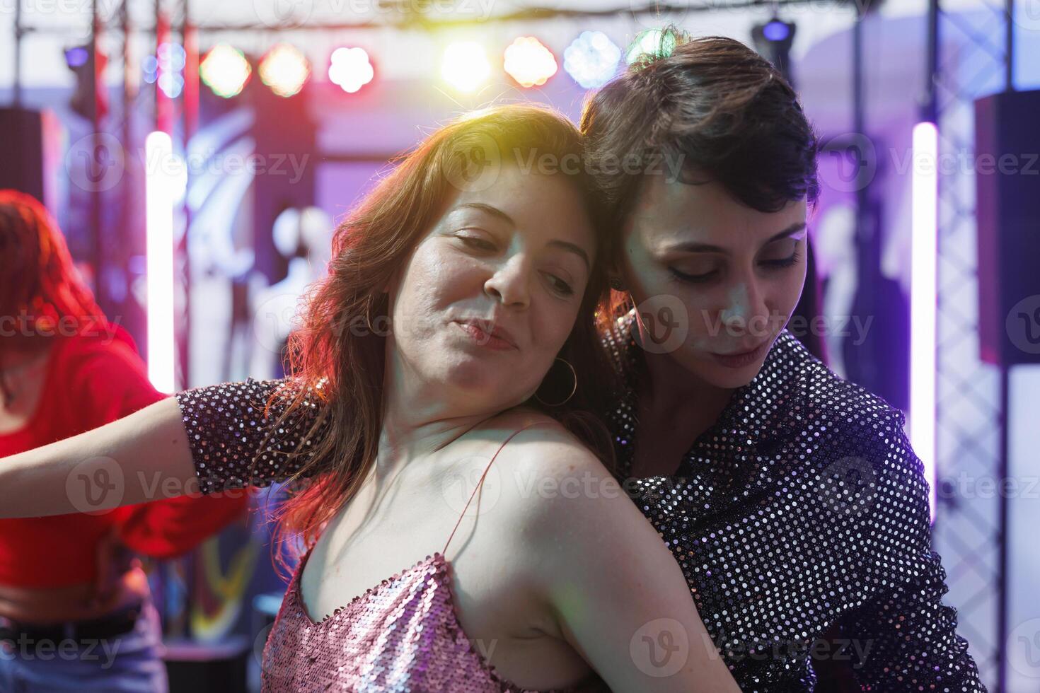Smiling women hugging while dancing together at nightclub party. Young romantic girlfriends couple relaxing and moving on crowded dancefloor while clubbing at discotheque photo