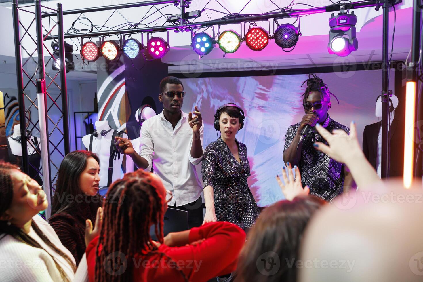 Band using electronic music digital controller and singing in microphone at nightclub disco party. Musicians playing on stage with spotlights while crowd moving on dancefloor photo