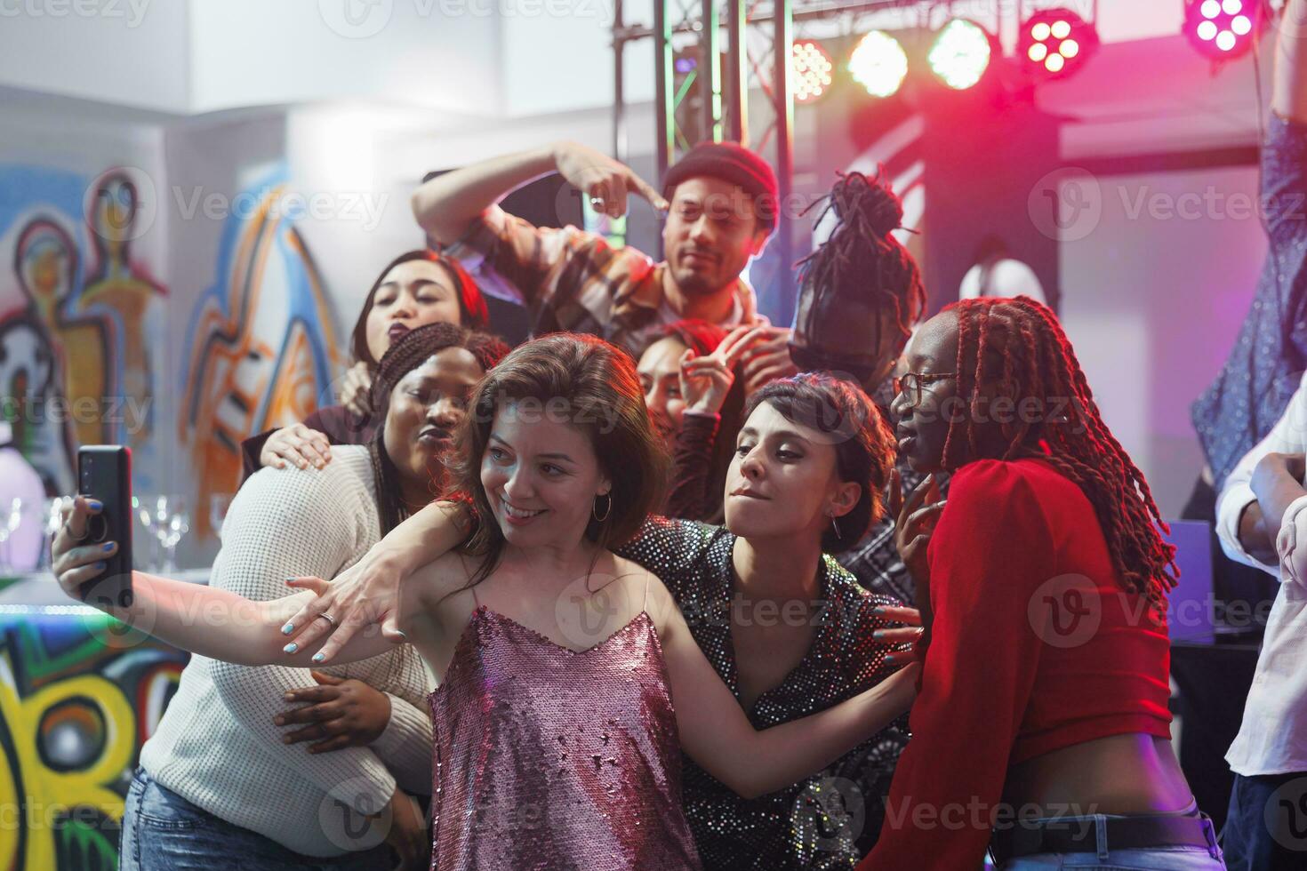 Group of friends posing for selfie on crowded nightclub dancefloor while clubbing together. Young carefree people taking photo on smartphone while enjoying discotheque event