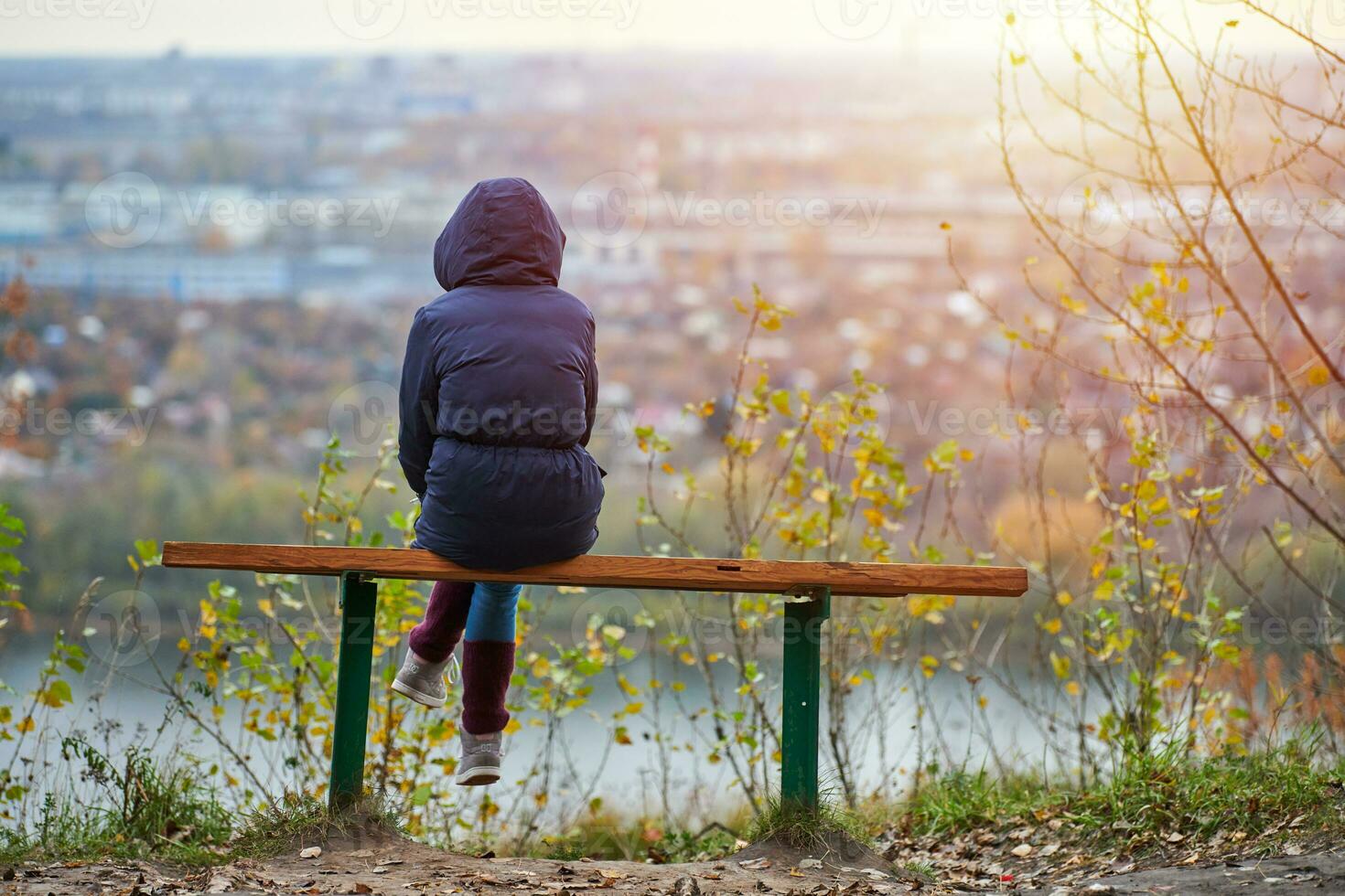 Young woman sitting on bench in autumn city park and looking at cityscape, back view photo