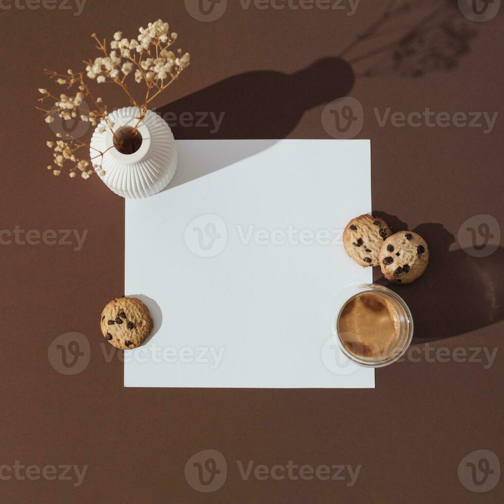 Creative trendy layout made with cup of coffee, chocolate cookies, white vase with dry flowers and paper card note copy space on espresso brown background. Minimal concept. Flat lay. photo