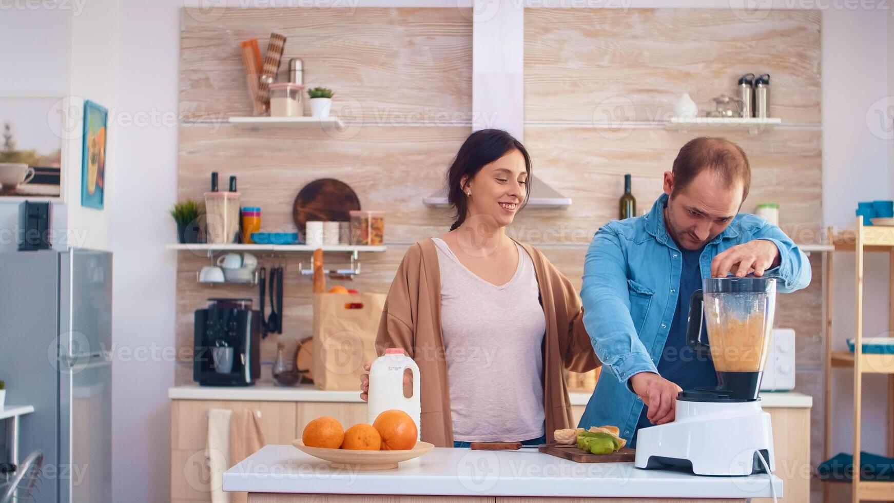 Man mixing fruits for smoothie using blender in kitchen. Cheerful young couple. Cheerful family making together organic healthy fresh nutritious tasty juice for breakfast from fresh fruits while on a diet. photo