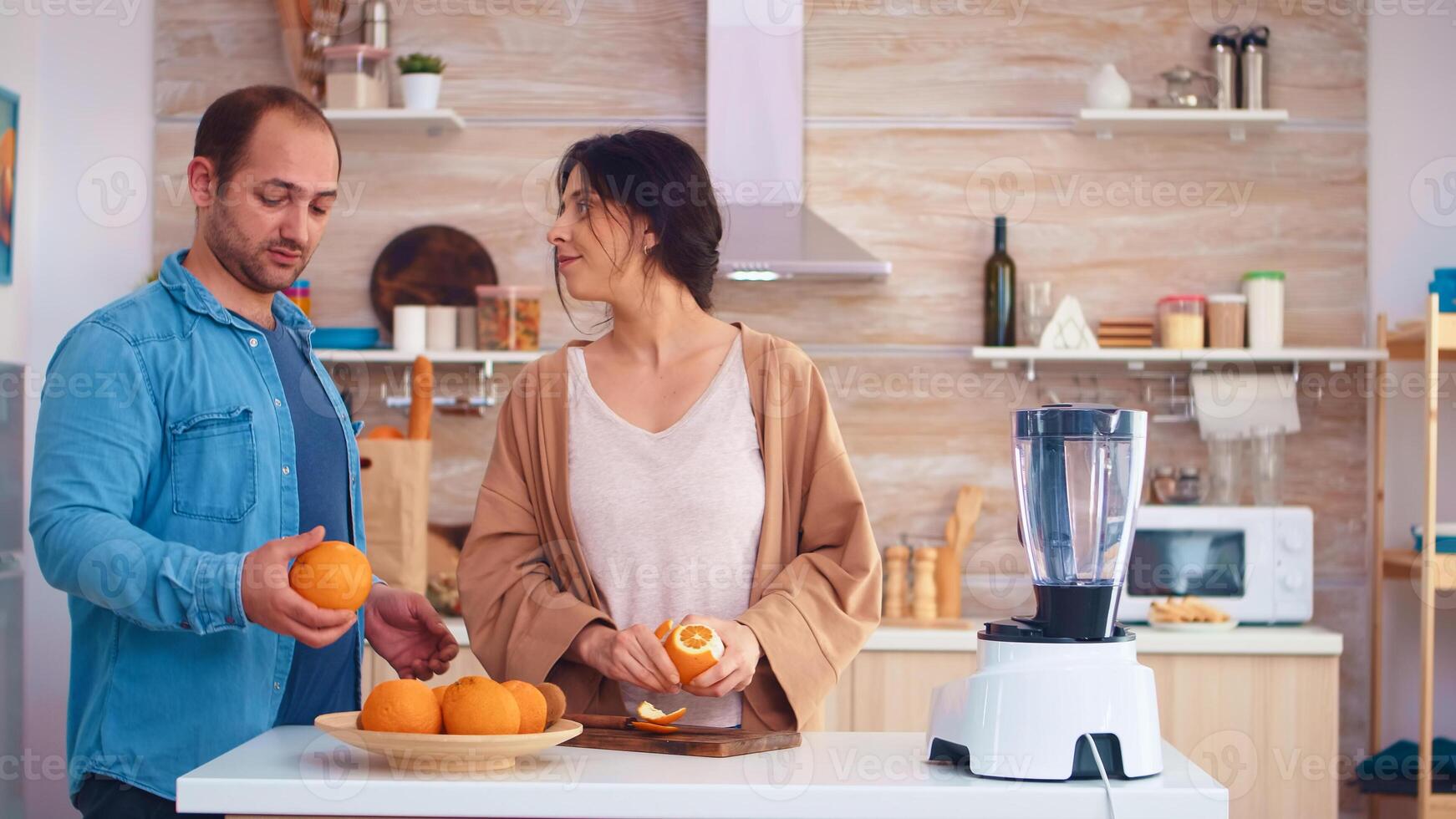 Couple making smoothie in kitchen. Woman peeling of orange while smiling at husband. Cheerful family making together organic healthy fresh nutritious tasty juice for breakfast from fresh fruits while on a diet. photo