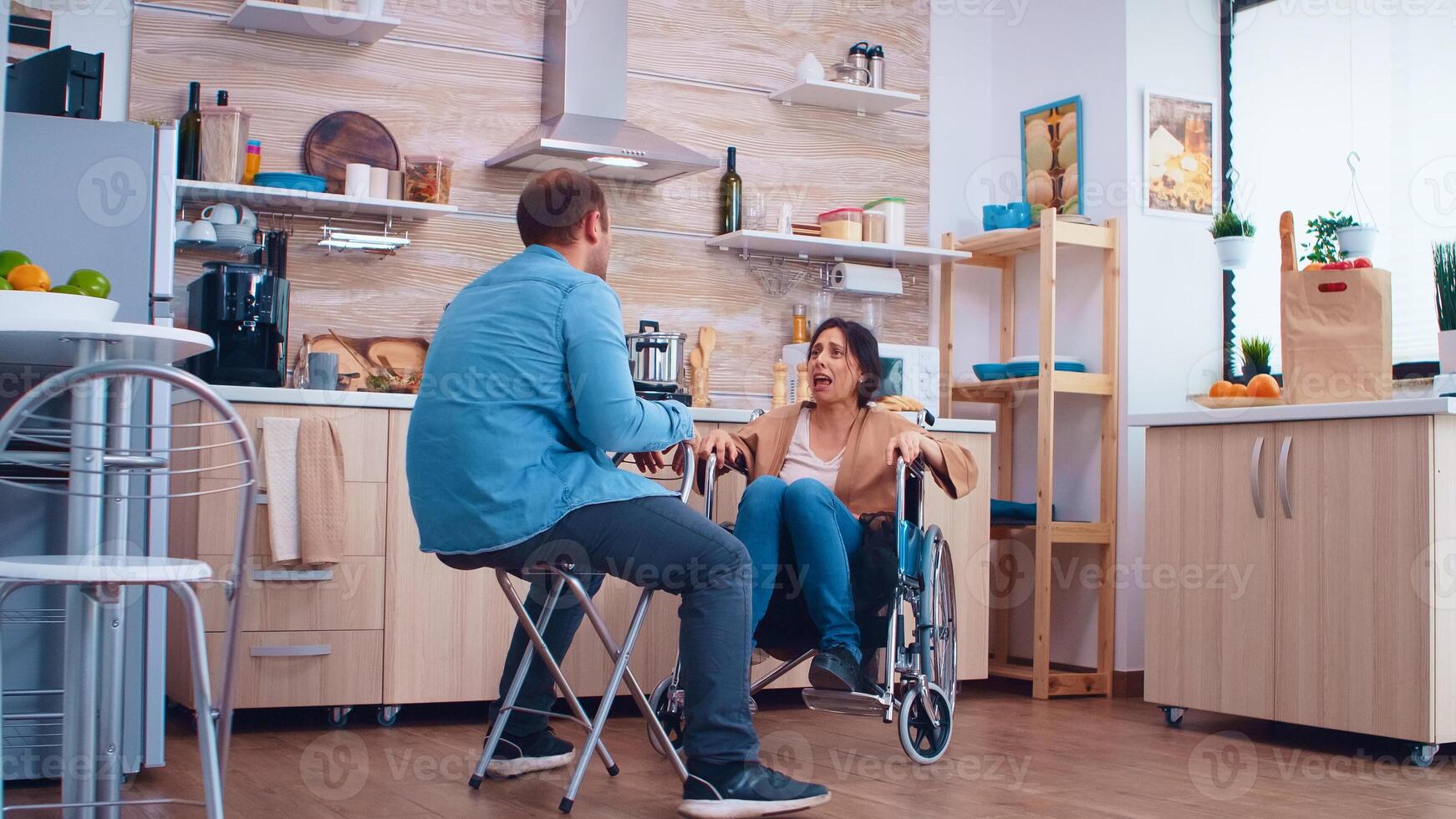 Unhappy disabled wife in wheelchair because of disagreement with husband in kitchen. Woman with paralysis handicap disability handicapped difficulties getting help for mobility from love and relationship photo