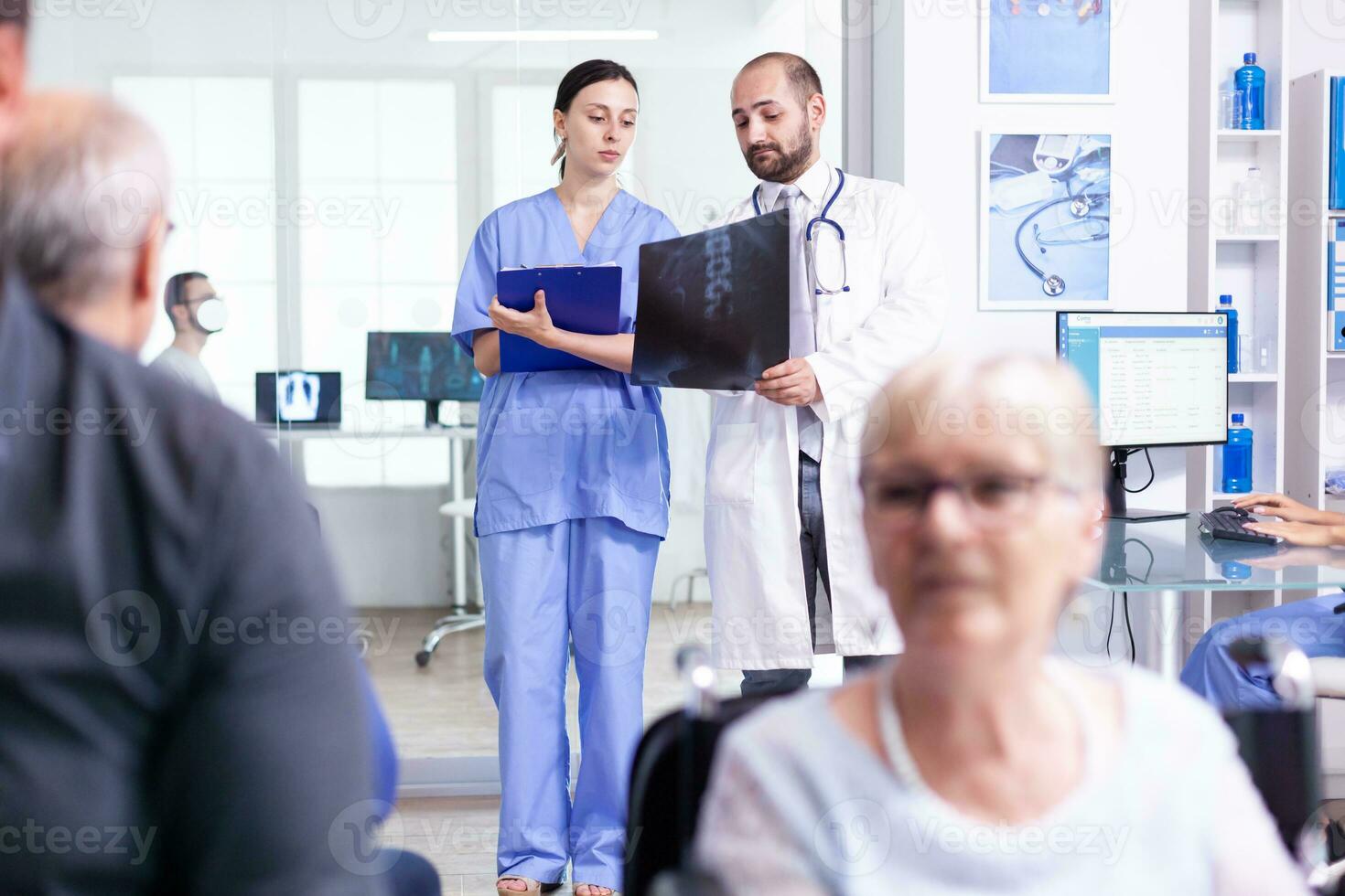 Nurse looking at radiography while talking with doctor about patient diagnosis in hospital waiting area. Invalid old woman in wheelchair. Senior man waiting for medical examination. photo