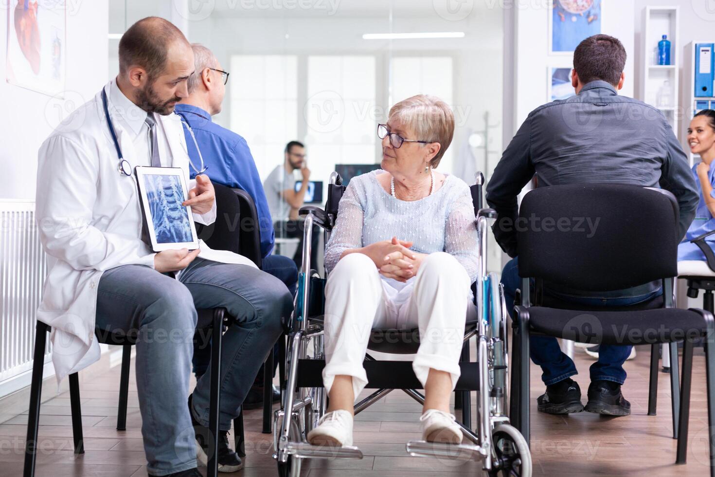 Doctor holding tablet pc with x-ray while explaining diagnosis to disabled elderly woman in wheelchair. Handicapped patient in hospital waiting area. Man in examination room. photo